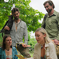 Four adults outdoors in lush greenery, wearing adventure or expedition clothing, two standing with gear like bows and arrows, two seated and reading a map or paper, suggesting a wilderness exploration or survival scenario.