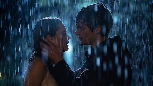 Two people stand close together in the rain at night, gazing into each other's eyes as one gently holds the other’s face, surrounded by falling water and dramatic lighting, creating a romantic and emotional atmosphere.