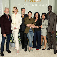 Bradley Whitford, Allison Janney, Rufus Sewell, Keri Russell, Debora Cahn, Ali Ahn and Ato Essandoh stand together posing for a group photo at an event for "The Diplomat," in front of a green and white decorative backdrop with Netflix branding.