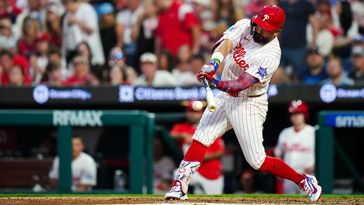 Philadelphia Phillies baseball player in white pinstripe uniform hitting the ball during a game, with a crowd of fans in the stadium background. Daytime, action shot at home plate.