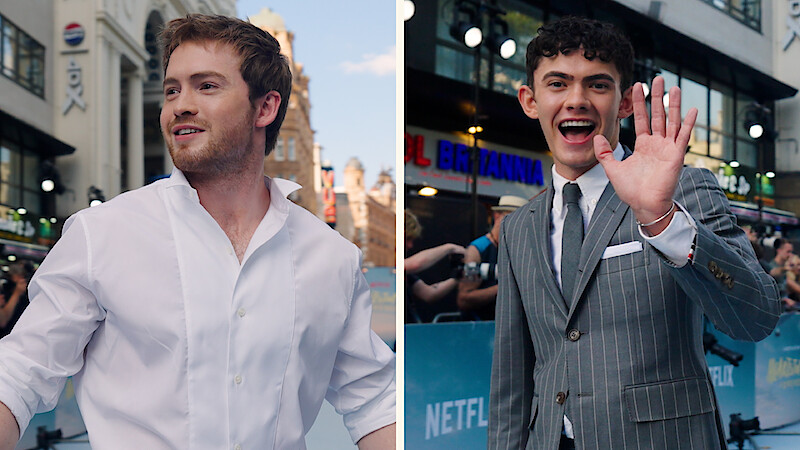 Kit Connor and Joe Locke at the Heartstopper Forever premiere, one in a white shirt smiling, the other in a pinstripe suit waving, with city buildings, lights, and Netflix banners in the background.