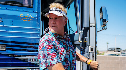 Man in a tropical shirt and visor stands at the doorway of a blue luxury bus on a sunny day, holding the door handle, with a fence and clear sky in the background.