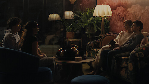 Four people sit across from each other in a dimly lit cozy living room with vintage furniture, lamps, houseplants, and a round coffee table, suggesting an intimate evening conversation.