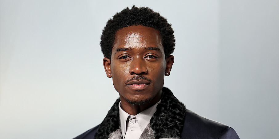 A well-dressed man with short curly hair and earrings standing in front of a plain, light-colored background, photographed in a studio setting with soft lighting.