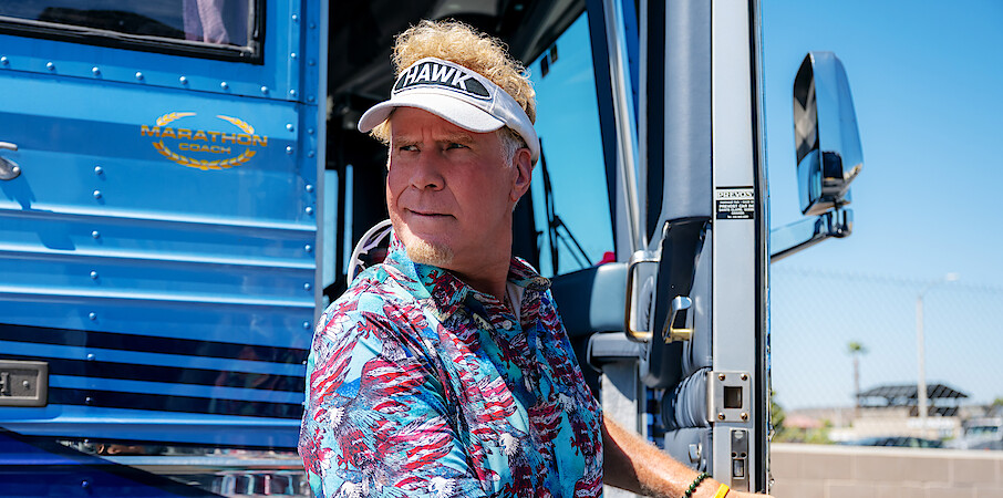 Man wearing a tropical shirt and visor stands at the open door of a blue RV in a sunny outdoor setting with a fence, palm trees, and blue sky in the background.