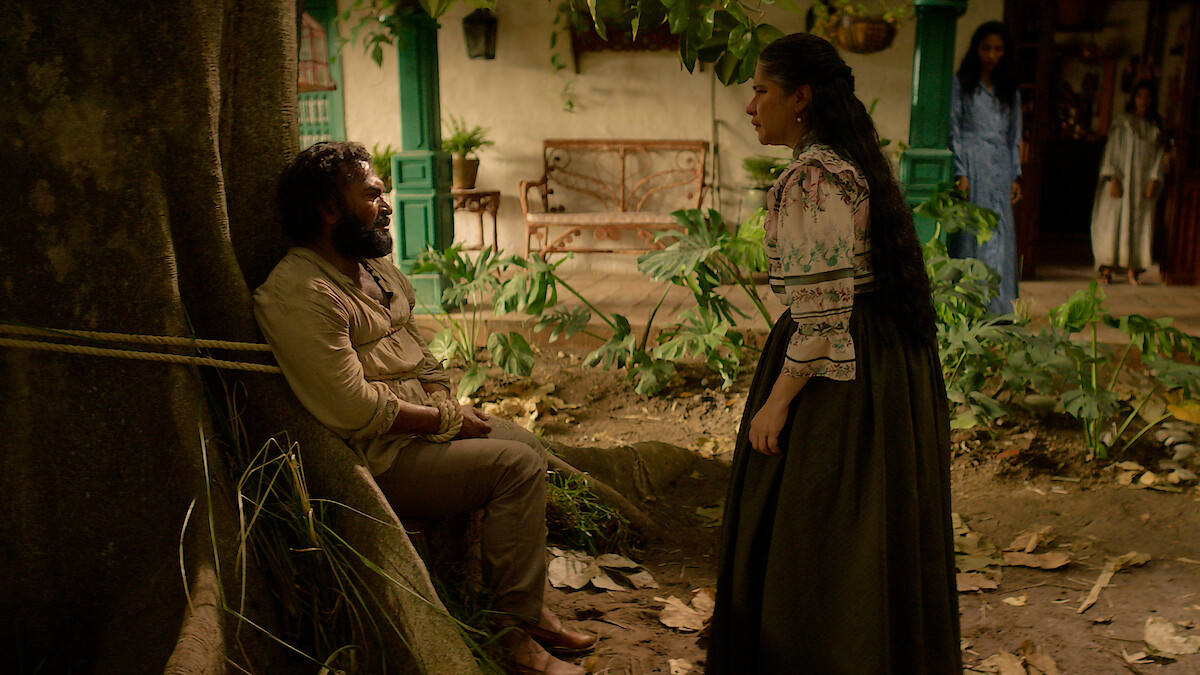 A man tied to a tree sits and faces a standing woman in 'One Hundred Years of Solitude'