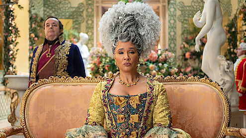 Woman in elaborate 18th-century costume and white wig sits on ornate gold sofa in a lavish, floral-decorated room; man in historical attire stands behind; statue and flowers in background.