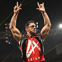 Muscular man wearing sunglasses, a vest, and a red shirt with "LA Knight" written, standing in an arena with bright lights, raising both arms and making hand gestures during what appears to be a wrestling event.