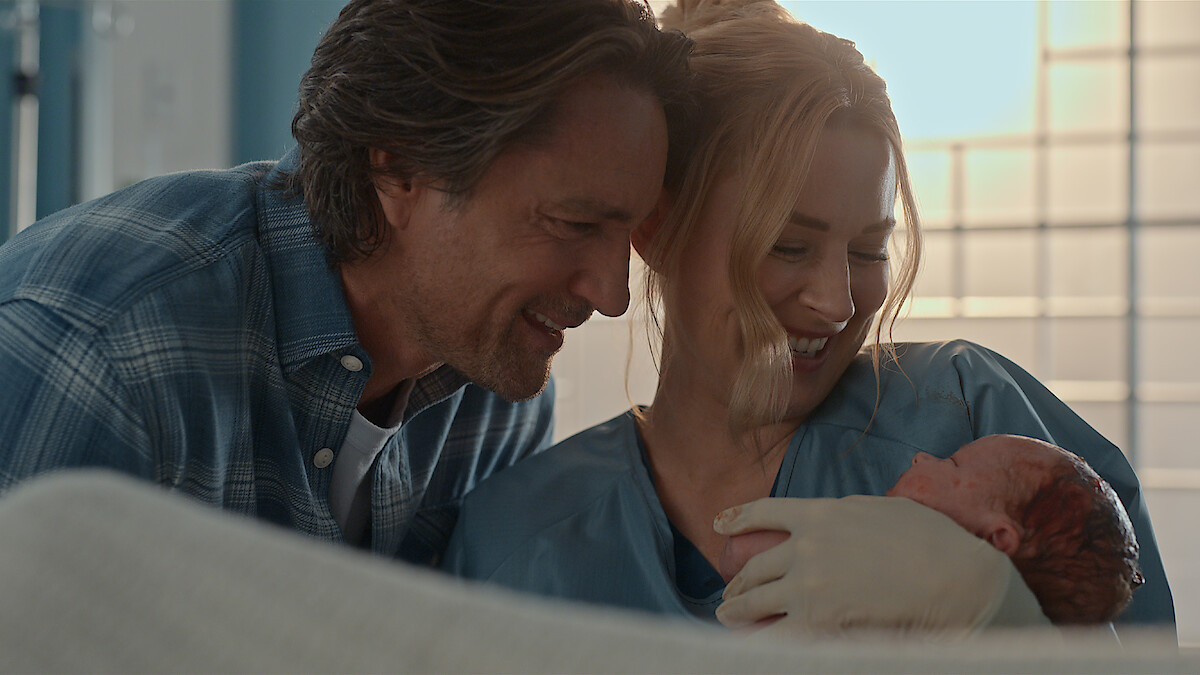 A happy couple in a hospital room lovingly cradle their newborn baby. The mother, in medical scrubs, smiles down at the baby while the father leans close, both appearing joyful and emotional in the bright, softly lit setting.