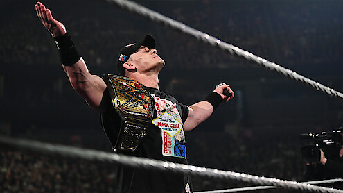 Wrestler in a black shirt and cap stands in a wrestling ring, holding a championship belt, arms raised triumphantly, with a cheering crowd in a large arena, creating an energetic and victorious atmosphere.