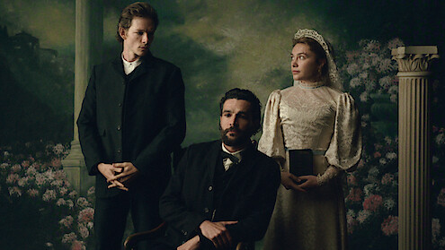 Three people in vintage clothing pose for a formal portrait with a scenic painted backdrop and decorative column, evoking a 19th-century setting. One woman in a lace dress and two men in dark suits.