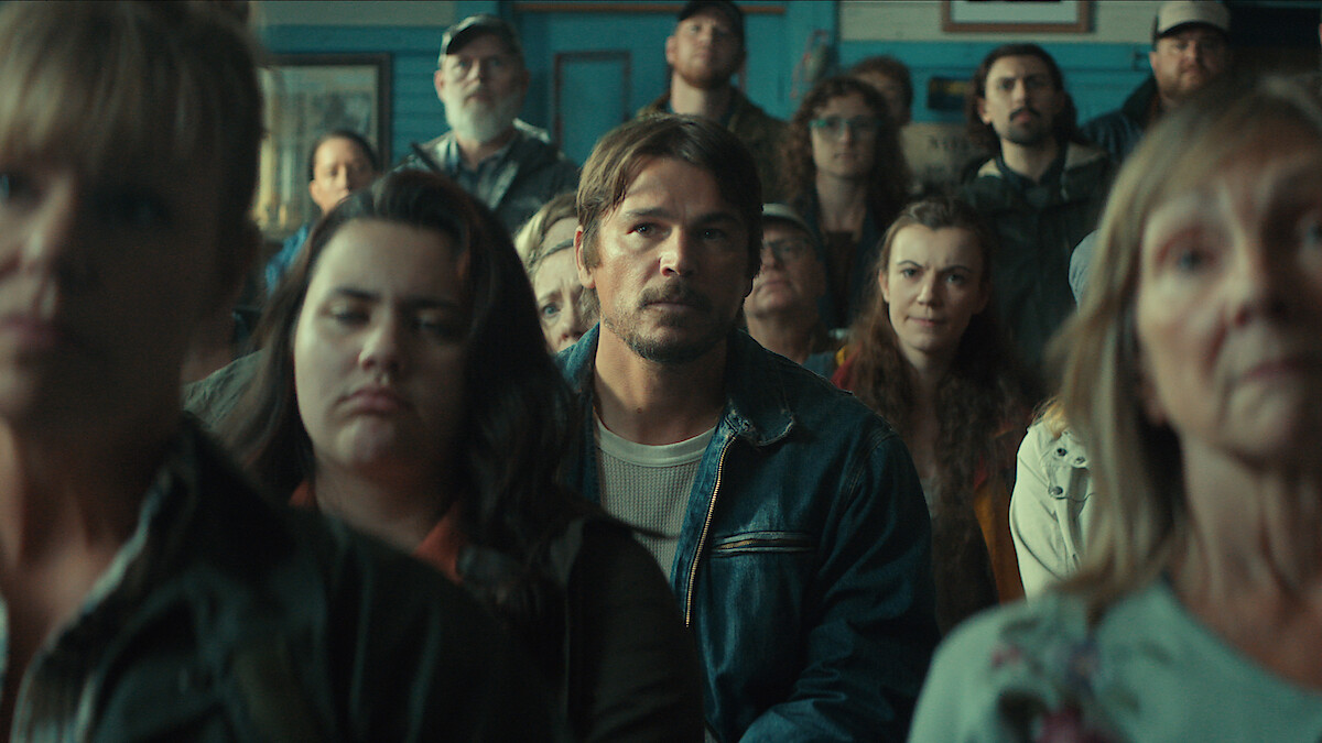 A group of people sit closely together indoors, appearing attentive and serious, likely at a community meeting in a rustic room with blue walls and casual clothing, with the central man in focus.