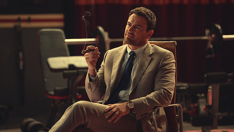 Man in a tan suit sitting in a chair indoors, holding a cigar and looking thoughtful. The background features gym or fitness equipment and dim, moody lighting.