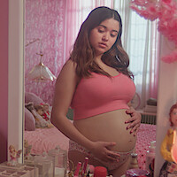 Pregnant woman in pink top and underwear stands in a pastel pink bedroom, looking down at her belly with hands on it, surrounded by toys, dolls, makeup, and soft light from a window with sheer curtains.