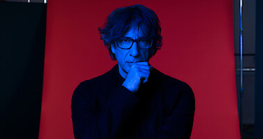 Neil Gaiman poses in front of a red seamless. 