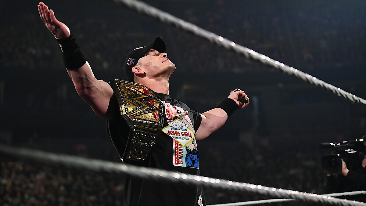 Wrestler in a black shirt and cap stands in a wrestling ring, holding a championship belt, arms raised triumphantly, with a cheering crowd in a large arena, creating an energetic and victorious atmosphere.