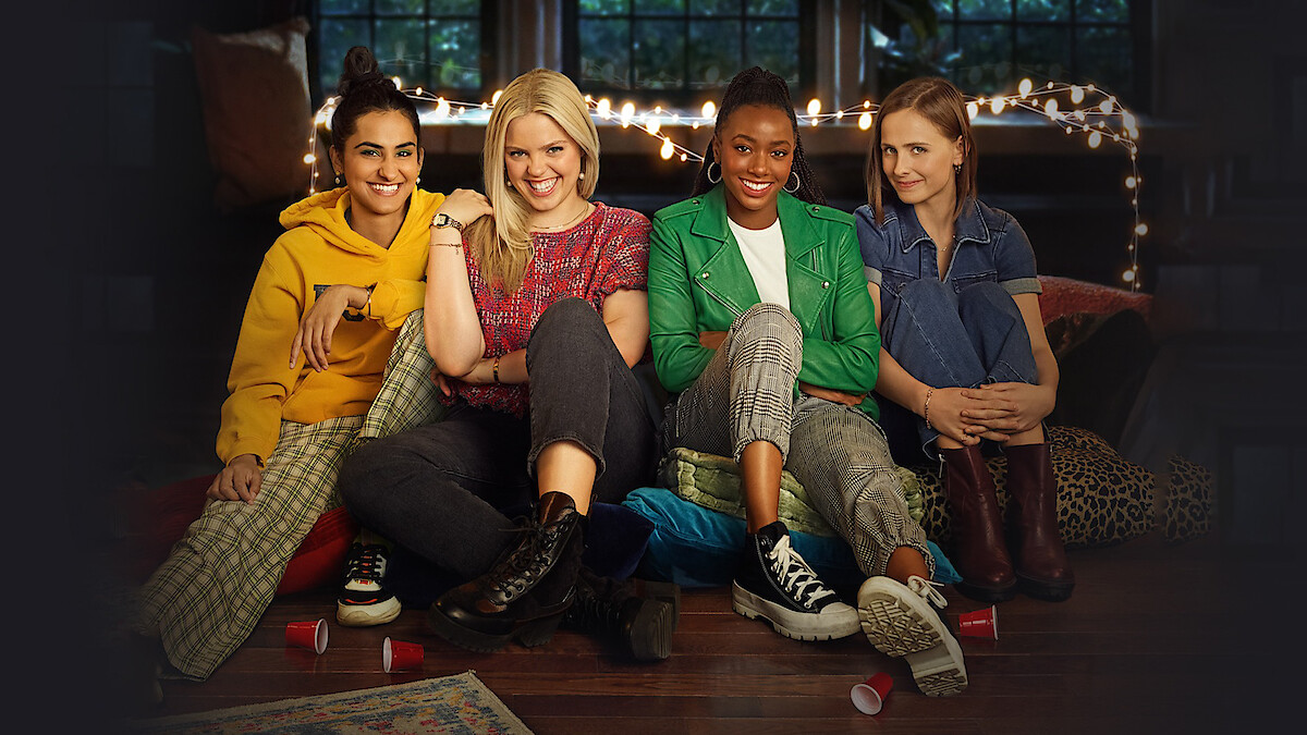 Four young women sit on the floor together in a cozy room with string lights and large windows, smiling and relaxed, surrounded by pillows and cups, suggesting a casual indoor gathering or hangout.