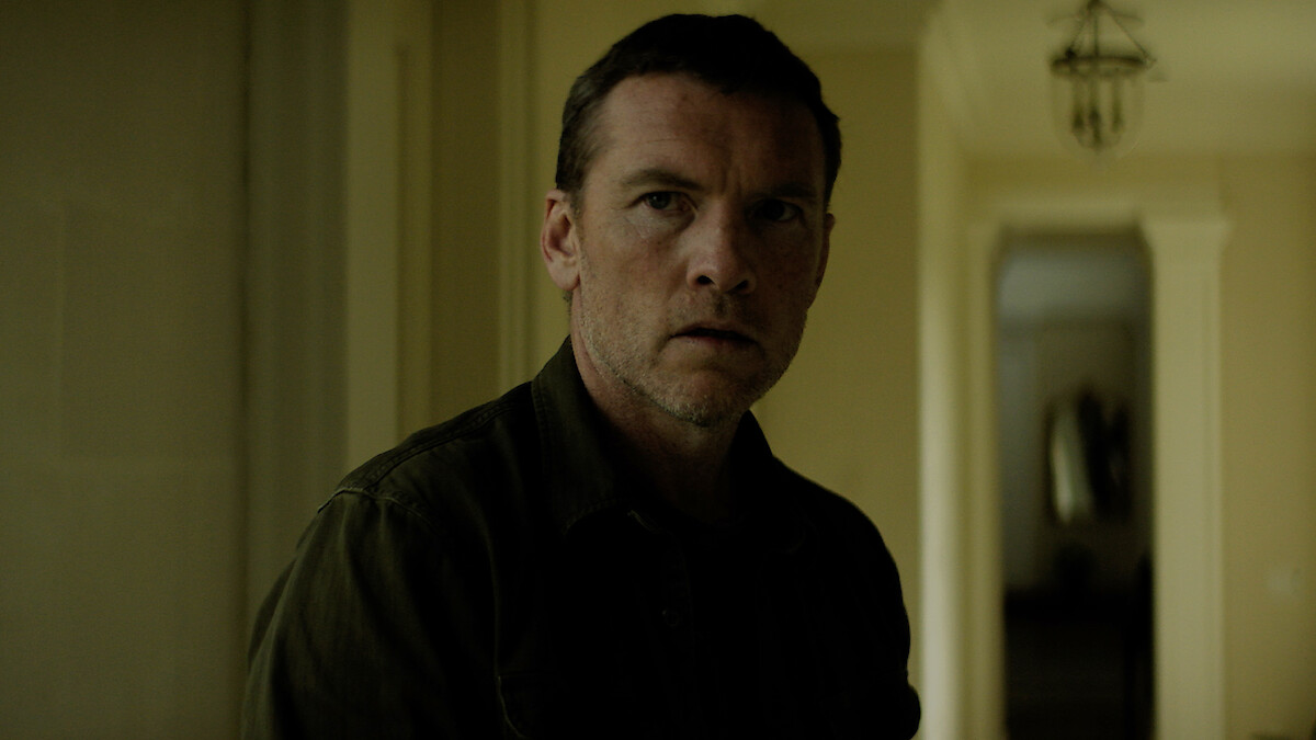 A serious man stands indoors in a dimly lit hallway with pale yellow walls and columns, looking slightly off-camera. The background features a corridor and a hanging light fixture.