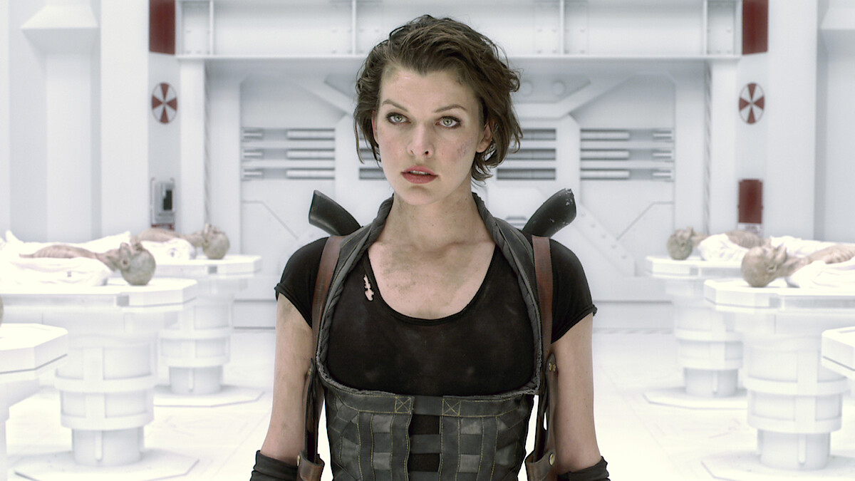 A woman in tactical gear stands in a futuristic, sterile laboratory with white walls and equipment, surrounded by examination tables holding covered bodies, suggesting a sci-fi or action setting.