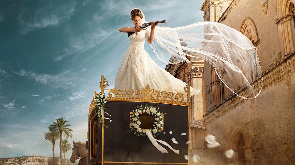 A bride in a white wedding dress stands on top of an ornate carriage, aiming a shotgun, with her veil flowing in the wind. The scene is set outdoors near classical buildings and palm trees under a bright sky.
