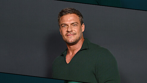Alan Ritchson with short hair wearing a dark green collared shirt standing against a plain dark background, facing forward and smiling slightly in a studio setting.
