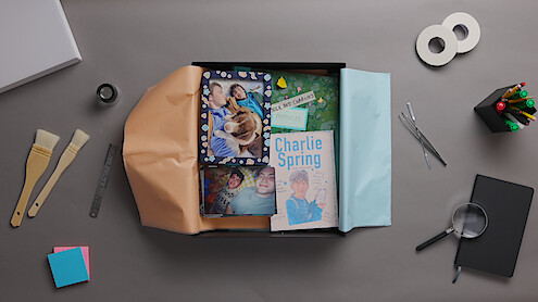 Scrapbook gift box with photos, book titled "Charlie Spring," and keepsakes on a desk. Surrounding items include paintbrushes, ruler, tape, pens, sticky notes, magnifying glass, scissors, and colored paper. Creative workspace setting.