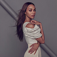 Zoe Saldaña stands against a gray background wearing an ensemble made of different shades of cream along with some gold jewelry, facing the camera with her hand touching her shoulder. 