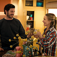 Two people smiling and exchanging mugs in a cozy kitchen, surrounded by yellow cabinets, flowers, and assorted kitchen items, suggesting a warm and casual atmosphere.