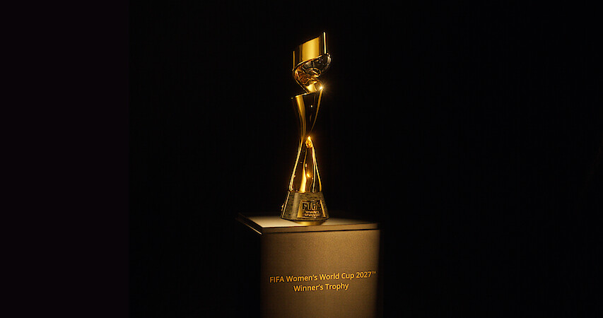 The image shows the FIFA Women’s World Cup 2023 winner’s trophy displayed on a pedestal in a dark environment, with focused lighting highlighting the shiny gold trophy and the engraved text on the base.