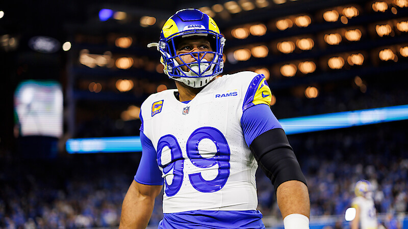 Football player in Los Angeles Rams uniform and helmet with number 99 stands on the field in a brightly lit stadium during a game, with a crowd visible in the background.