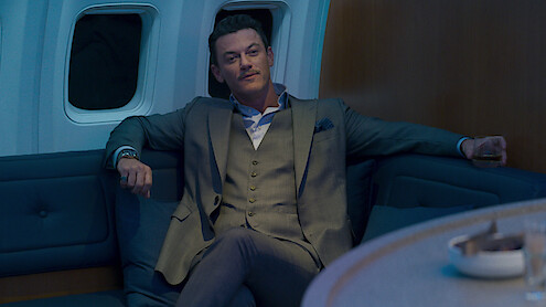 A man in a suit sits relaxed on a blue sofa inside a plane cabin, holding a drink with an arm resting on the seat, in a dimly lit, modern and luxurious environment.