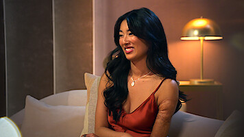 Emma Betsinger wearing a satin dress sits on a beige sofa in a softly lit, elegant room with a modern gold lamp and neutral-toned walls in the background.