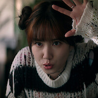 Young woman with brown hair in space buns, wearing a cozy striped sweater, making a playful gesture with her hand raised indoors, background softly blurred with plants and neutral tones.