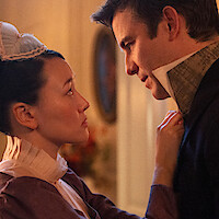 A woman in period costume faces a man in a formal suit in a softly lit, elegant Victorian-era room, with intense expressions suggesting a serious or intimate conversation.