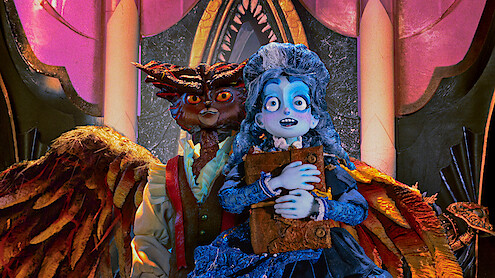 A blue-skinned, animated girl in a blue dress holds a book, standing nervously in front of a menacing owl-like creature with wings inside a lavish, ornately decorated room with pink and gold arches and dramatic lighting.