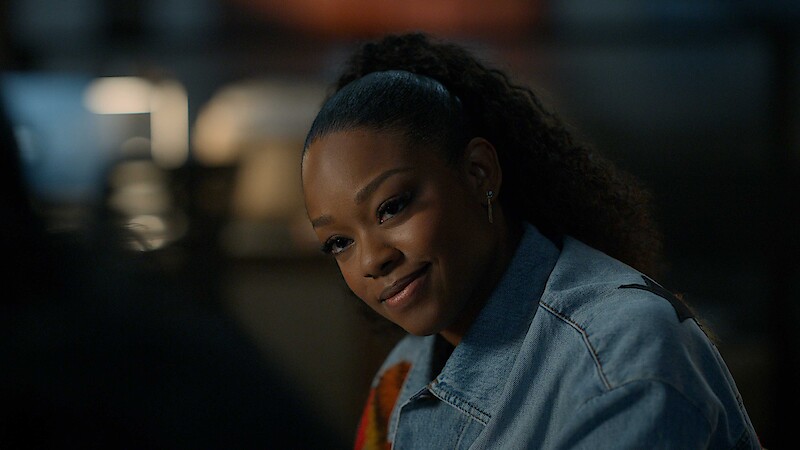 A woman with curly hair tied back is sitting indoors, wearing a denim jacket, and smiling warmly in a softly lit environment with a blurred background.