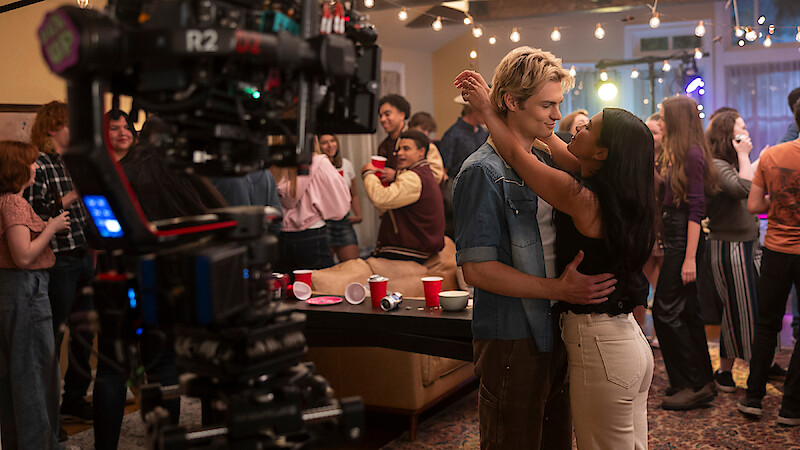 A film crew captures a young couple dancing closely at a lively indoor party, surrounded by friends, string lights, and festive decorations in a cozy living room setting.