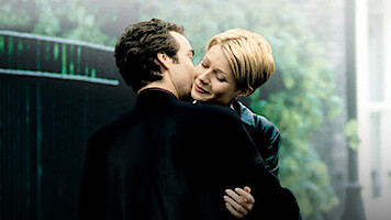 A man and woman embrace outdoors near a garden gate, with greenery and soft lighting in the background, conveying a romantic and intimate atmosphere.