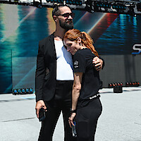 Two people embrace in a staged outdoor arena with SummerSlam and WWE logos in the background, under bright sunlight, conveying a calm and supportive mood before a wrestling event.