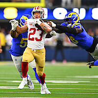 Football player in red and gold uniform runs with the ball on a stadium field, pursued by two defenders in blue and yellow uniforms during an NFL game.