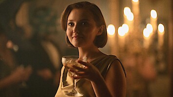 Woman in a gold dress holding a drink at an elegant, dimly lit party with candlelight in the background, creating a warm, sophisticated atmosphere.