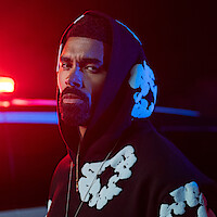 Man in black hoodie with white floral patterns stands in front of a police car with bright red and blue lights at night, looking serious.