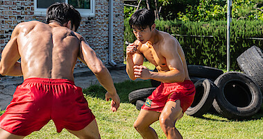Two muscular  boxers in red shorts square off on a lawn with old tires stacked up behind them. 