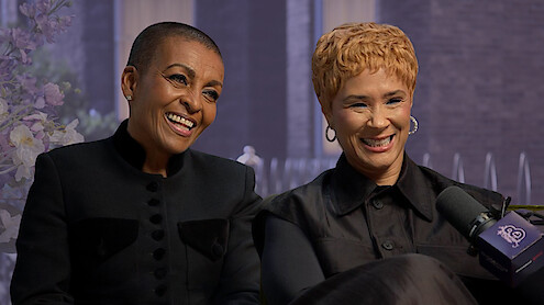 Two women smiling during an interview, seated indoors with a floral arrangement and large windows in the background, both wearing black outfits, with a microphone labeled “20” visible in the foreground.