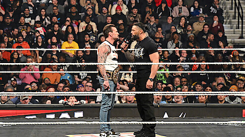 Two wrestlers face off in a packed arena wrestling ring, one holding a championship belt and the other speaking into a microphone as a large audience watches the dramatic confrontation.