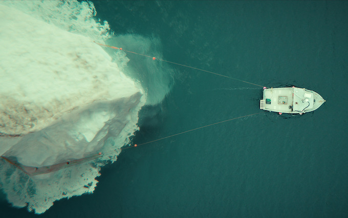 Calvin tows the iceberg through deep water in Below An overhead view shows a small fishing boat towing an iceberg across dark ocean water in Netflix Below