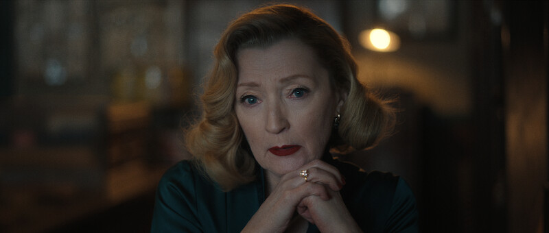 A woman with styled blonde hair and red lipstick sits with her hands clasped under her chin, looking thoughtfully ahead in a softly lit room.