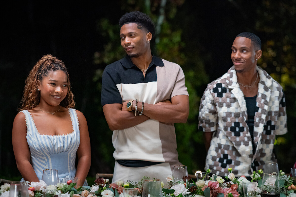 Three people standing behind a decorated dinner table outdoors at night, dressed in stylish casual attire, surrounded by flowers and candles, appearing to enjoy a social gathering or celebration.