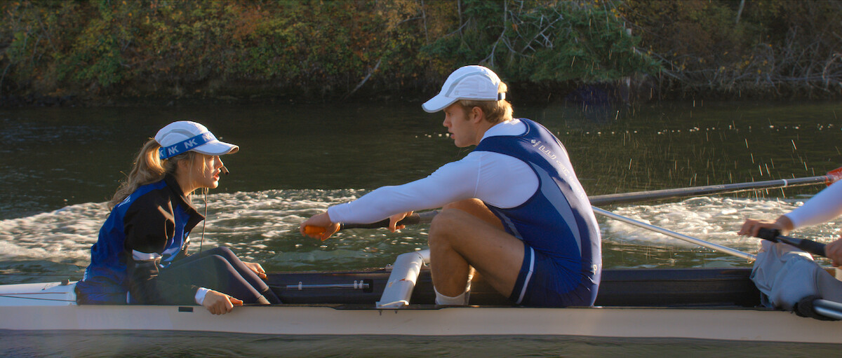 Rowers in blue and white uniforms compete on a calm river, with a female coxswain guiding the boat and trees in the background, sunlight reflecting off the water.