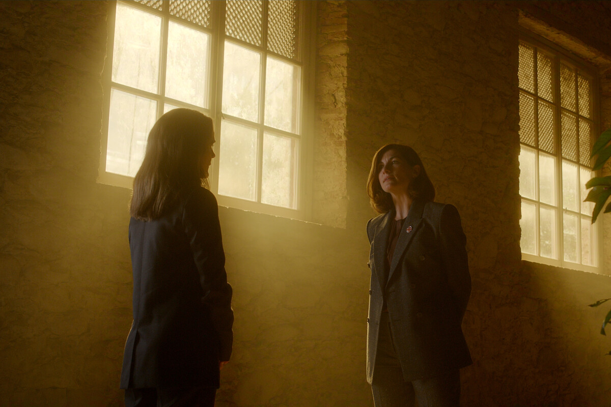 Two people in suits stand facing each other in a warmly lit room with large windows and stone walls, sunlight streaming in, creating a dramatic atmosphere.